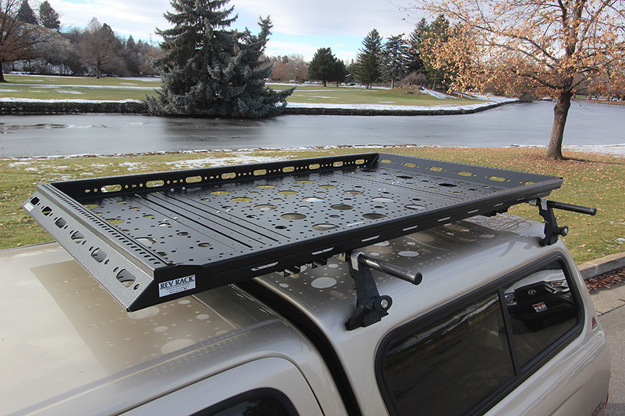 a medium sized black rev rack tray atop a gold toyota tacoma with gold topper shell, with a pond and trees in the background
