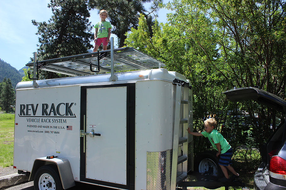 looking up at a child standing atop a large grey revrack tray attached to a enclosed white trailer in a campground setting