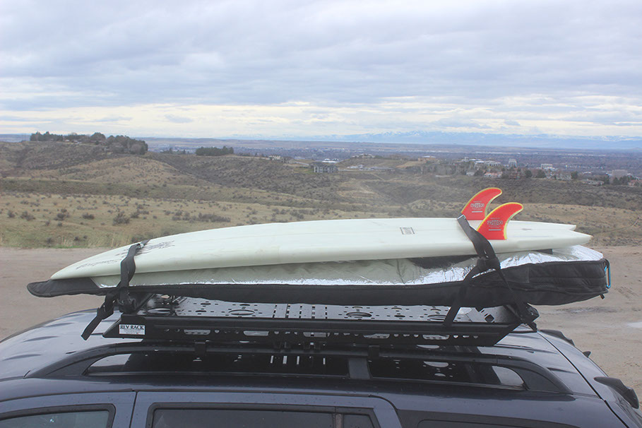 Two surfboards, one in a black bag and the other uncovered, white with 2 red fins, strapped to a small black rev rack tray atop a hun die 