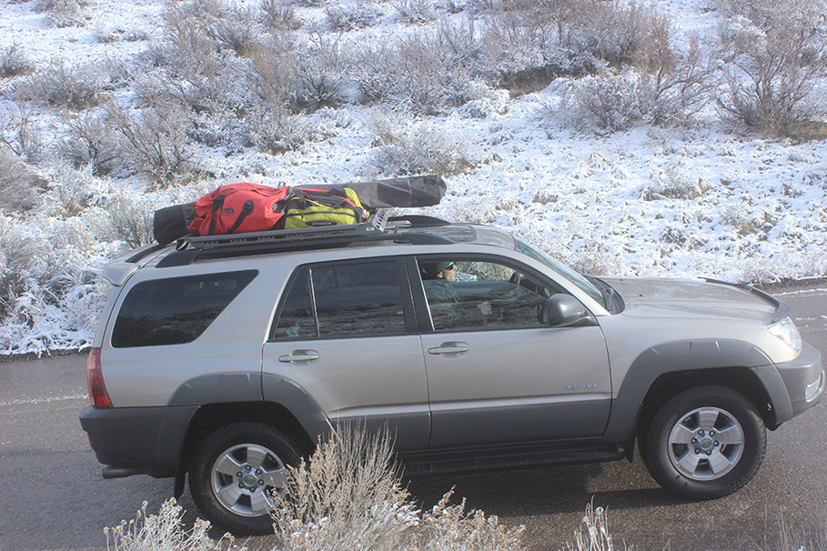 profile of a passing gold nissan pathfinder with a small revrack tray on top and winter gear including snowboards with a snowy background