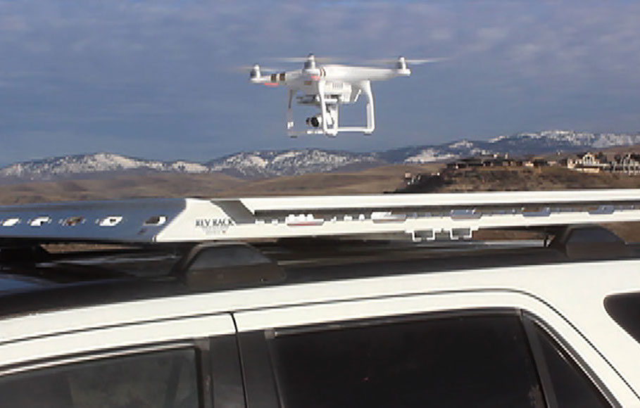 profile view of a white drone hovering above a small white revrack tray atop a white honda c r v with a foothill background